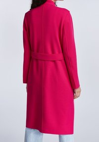 Fuksiankeltainen villatakki, jossa on pitkät hihat, vyötäröllä vyö ja takana halkio. Saumalistausta korostaa rakennetta ja muotoilua.