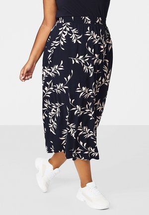 Kobieta w czarnej spódnicy midi w białe liście i białych tenisówkach, idąca na tle jasnego, jednolitego tła.