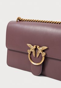 Bolsa de ombro de pele roxa com corrente torcida dourada e fecho dourado, apresentando duas aves a enfrentar-se.