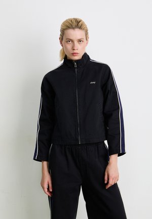 Svart cropped jakke laget av robust stoff, med høy krage, glidelås og kontrasterende hvite striper langs ermene.