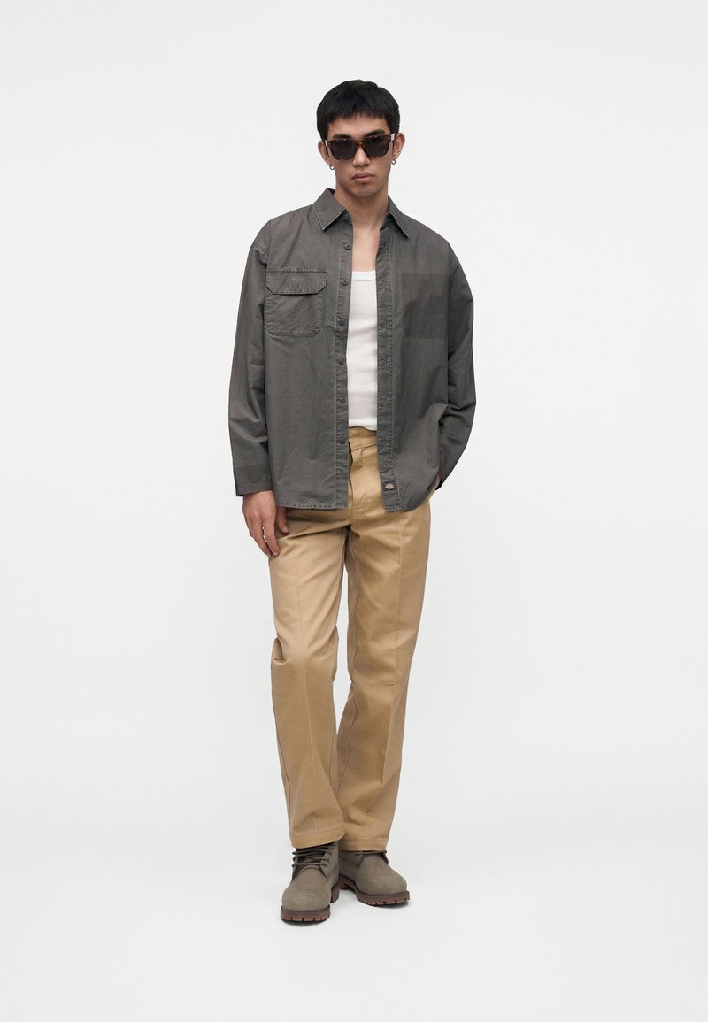 Uomo che indossa una camicia oversize grigio scuro abbottonata sopra una canottiera bianca, pantaloni beige, stivali marroni e occhiali da sole, in piedi davanti a uno sfondo bianco.