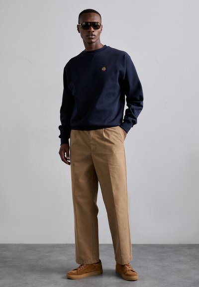 Navy sweatshirt with a small colorful logo, khaki straight-fit pants, and brown sneakers. Sleek sunglasses complete the outfit.