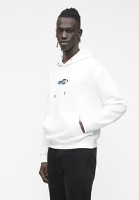 Jovem com cabelo escuro em tranças, vestindo um casaco branco com capuz com o logótipo "GUESS JEANS" e calças pretas, em pé com uma mão no bolso.