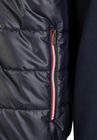 Marineblauw gewatteerd jack met een ritszakje met rode en witte accenten. De stof is glanzend, glad en doorgestikt voor isolatie.