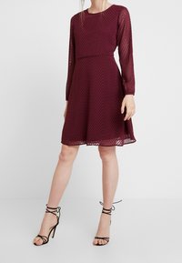 Burgunderfarbenes Kleid mit langen, durchsichtigen Ärmeln, das ein Zickzackmuster aufweist, einen taillierten Oberteil und einen ausgestellten Rock hat; kombiniert mit schwarzen Riemchen-Heels.