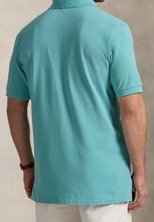 Mann von hinten, trägt ein türkisfarbenes kurzärmliges Poloshirt und weiße Hosen, steht vor neutralem Hintergrund.