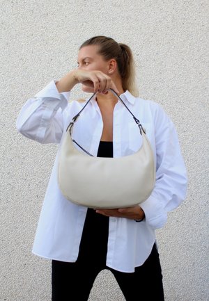 Cremefarvet læderhåndtaske med en buet form, der har en mørk rem. Modellen bærer en hvid skjorte med knapper og en sort top, mens hun poserer foran en tekstureret væg.