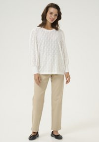 Pull blanc à motifs avec un design en trous en losange, manches ballons, associé à un pantalon beige et des chaussures plates noires, sur un fond uni.