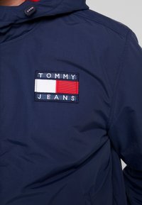 Marinejacke mit einem Stoffpatch mit "TOMMY JEANS" und einem rechteckigen Logo in Rot, Weiß und Blau. Glatte Textur, verstellbare Kapuze.