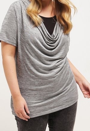 Femme portant un haut gris drapé à col bénitier et manches courtes sur un débardeur noir avec un jean foncé, debout devant un fond uni.