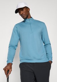 Un hanorac fermoar de culoare albastru deschis, cu guler înalt și mâneci lungi, purtat peste pantaloni închiși la culoare. Modelul ține un club de golf. O șapcă albă adaugă contrast.