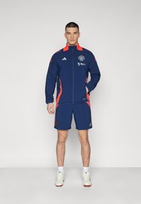 Tuta sportivo blu con dettagli rossi, composto da una giacca con zip e pantaloni corti abbinati. Include il logo della squadra e il branding dello sponsor.