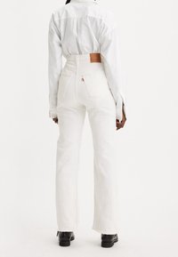 Jeans blancos de tiro alto con piernas anchas, que presentan bolsillos traseros y un parche de cuero marrón en la cinturilla. Botas negras completan el look.