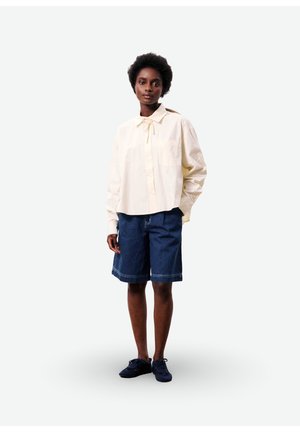 Vrouw staand, gekleed in een roomkleurige blouse met lange mouwen en een nektie, denim knielange shorts en zwarte sneakers tegen een witte achtergrond.