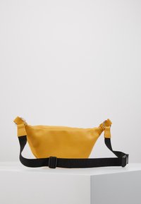 Marsupio sintetico giallo con texture liscia, tracolla regolabile nera e accessori in argento. Forma curva unica e design minimalista.