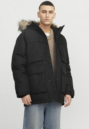 Sort isoleret parka-jakke med hætte pyntet med pels, flere frontlommer og lynlåsclosure. Bærer over en beige sweater og jeans.