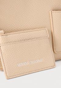 Šviesiai rudos tekstūruotos odinės kortelių laikiklis su matomomis siūlėmis ir įspaustu "ARMANI EXCHANGE" logotipu. Plonas dizainas su dviem kortelių lizdais.