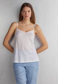 Witte, lichte cami met dunne bandjes en een V-hals. Zachte textuur en iets doorzichtige stof. Gepaard met lichtblauwe broek.