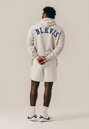 Hombre de pie de espaldas, vistiendo una sudadera con capucha gris con el texto "BLKVIS", pantalones cortos blancos con rayas finas, zapatillas blancas Nike y gafas, con las manos entrelazadas detrás de la espalda.