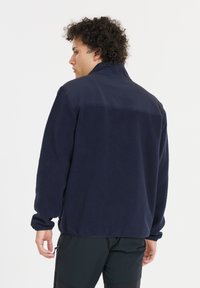 Marinblå fleecejacka med hög krage, texturerad tyg och slät axelpanel. Har långa ärmar och en avslappnad passform.