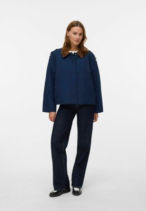 Giacca impermeabile blu navy con colletto marrone, tasche frontali e dettagli sulle spalle. Abbinata a jeans scuri e scarpe nere.