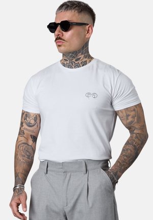 Mann mit Tattoos und Sonnenbrille, der ein weißes T-Shirt und graue Hose trägt, steht mit einer Hand in der Tasche vor einem einfarbigen Hintergrund.