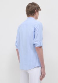 Camicia a righe blu chiaro con maniche arrotolate e colletto con bottoni. Il retro presenta un orlo curvo e una consistenza morbida.