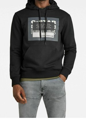 Mężczyzna ubrany w czarną bluzę z kapturem G-Star Raw z przednią kieszenią kangura i jasnoszare jeansy, stoi z jedną ręką w kieszeni.