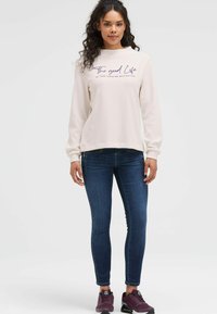 Helles beiges Rundhals-Sweatshirt mit dem Text "The good Life", kombiniert mit dunkelblauen Slim-Fit-Jeans und lila Sneakers.