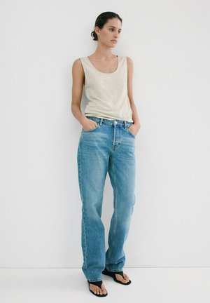 Žena stojaca opretá o bielu stenu, oblečená v bezrukávovom béžovom topánke, voľných modrých džínsoch a čiernych sandáloch s tenkými remienkami, s rukami v vreckách.