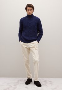 Navy blue cable-knit turtleneck sweater, cream-colored pants, and dark shoes. Model stands with hands in pockets against a neutral background.