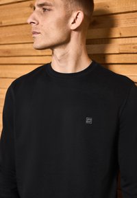 Schwarzes Langarmshirt aus glattem Stoff mit einem kleinen grauen Logo auf der Brust, vor einem Hintergrund mit Holzstruktur.