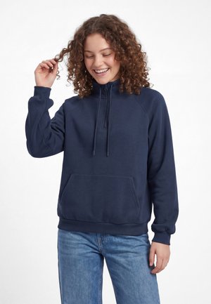 Ung person med krøllet hår, der smiler, mens de holder en hårtot, iført en marineblå kvart-lynlåstrøje og blå jeans mod en hvid baggrund.