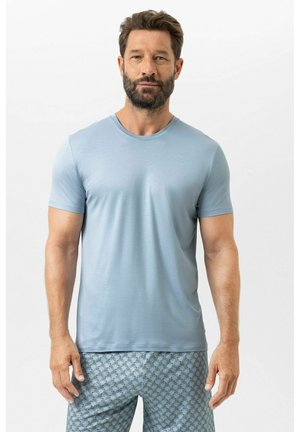 Mann mit Bart, der ein schlichtes hellblaues kurzärmeliges Hemd und gemusterte blaue Shorts trägt, steht vor weißem Hintergrund.