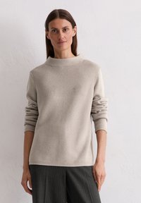 Beige Strickpullover mit hoch geschlossenem Kragen und weiten Ärmeln. Verfügt über ein strukturiertes Muster. Getragen mit grauen Nadelstreifen-Hosen.