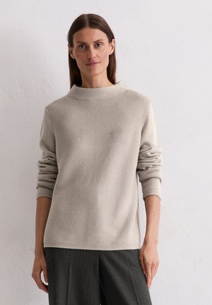 Maglione beige a maglia con collo alto e maniche larghe. Presenta un motivo testurizzato. Indossato con pantaloni grigi a righe.