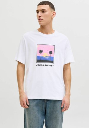 Junger Mann trägt ein weißes T-Shirt mit einem pink-blauen Sonnenuntergang und Palmenmotiv über dem Schriftzug "Jack & Jones", kombiniert mit blauen Jeans.