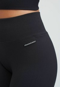 Leggings neri a coste con vita alta e una piccola etichetta con logo sull'anca sinistra. La texture è liscia con linee verticali prominenti.