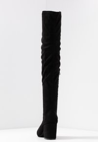 Stivale alto in suede nero con tacco a blocco e zip laterale, presentato su uno sfondo bianco con leggero ombreggiamento.