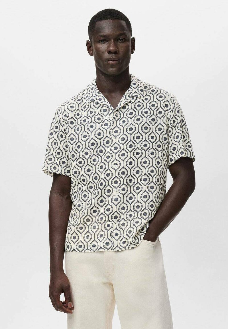 Camisa de manga corta con un patrón geométrico en azul marino y crema, con un collar doblado y una tela suave. Combinada con pantalones de color claro.