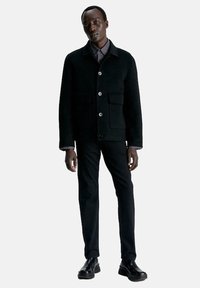 Zwarte jas van een wolmix met knopen aan de voorkant, twee borstzakken en een kraag. Gecombineerd met zwarte slim-fit broek en glanzende zwarte laarzen.