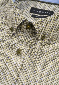 Beige Kurzarmhemd mit gemusterten kleinen Quadraten in Blau und Gelb. Verfügt über braune Knöpfe und einen hellblauen Innenkragen.