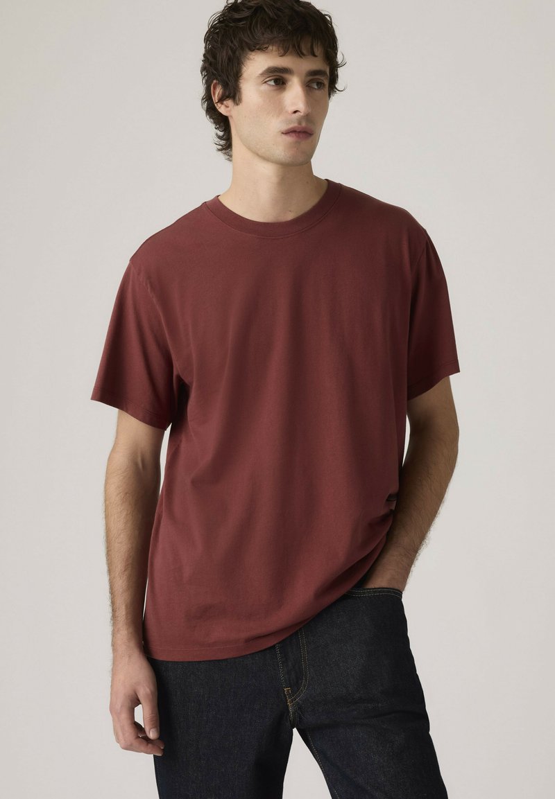 Giovane uomo con capelli ricci scuri che indossa una maglietta a maniche corte color amaranto semplice e jeans in denim scuro, in piedi con una mano in tasca.