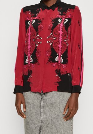 Blouse rouge à manches longues avec col et poignets noirs, ornée de motifs symétriques roses et noirs et de détails en forme d'éclairs sur les manches.