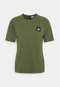 Skrócony rękaw, oliwkowa koszulka z miękkiego materiału, z czarną kwadratową naszywką z białym brandingiem Adidas na lewej stronie klatki piersiowej.
