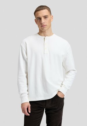 Giovane uomo con i capelli corti che indossa una maglietta henley bianca a maniche lunghe e pantaloni scuri, in piedi con una mano in tasca.