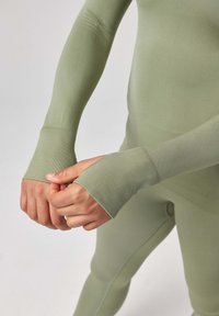 Langärmliges, enganliegendes Oberteil aus muted grünem Material mit Daumenlöchern an den Bündchen. Glatte Textur und keine sichtbaren Muster oder Verzierungen.