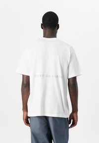 T-shirt en coton blanc avec une coupe décontractée, affichant "PAIX SUR TERRE" en texte gris clair au dos, encolure ronde et manches courtes.