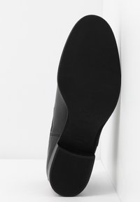 Chaussure en cuir noir avec une texture lisse et un bout arrondi. Dispose d'une semelle en caoutchouc plate avec un motif rainuré pour une meilleure adhérence. Marquée du nom de la marque.