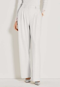 Vrouw in hoge taille, wijde witte broek gecombineerd met een witte geribbelde trui met kantenkraag en zilveren puntige hakken.